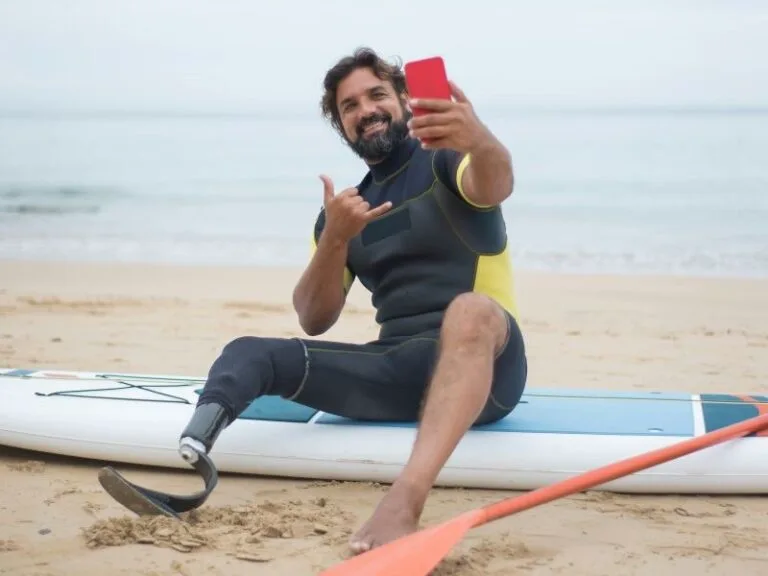 Man with prosthetic leg taking selfie on stand-up paddle board at beach.