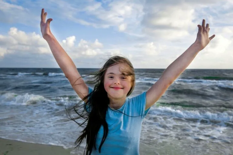young girl with down syndrome smiling with her hands up in the air with the beach as a backdrop