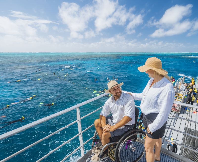 Man in wheelchair and woman overlooking snorkellers in blue water.