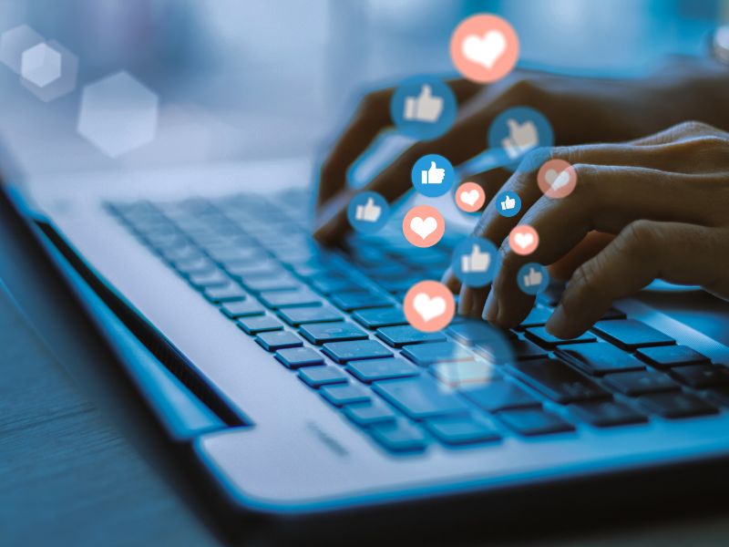How to write social media captions for tourism business - hads typing on a keyboard. graphics of social media 'likes' and 'hearts' overlaid on top