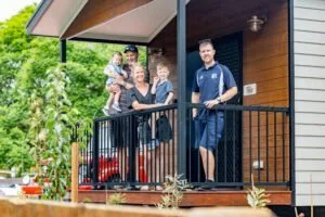 Family of five standing on cabin deck with green gardens and red outdoor furniture visible.