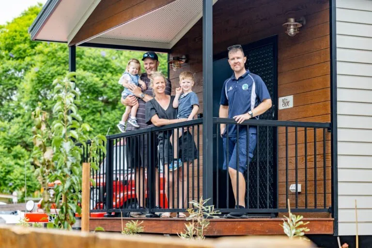 Family of five standing on cabin deck with green gardens and red outdoor furniture visible.