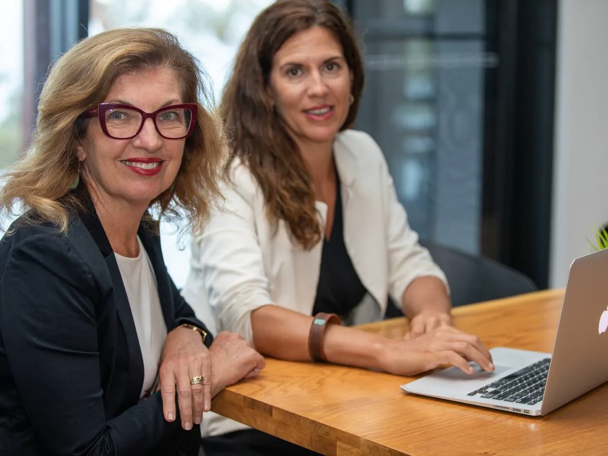 Fab and Liz sit at a wooden desk with a laptop, providing digital marketing support.