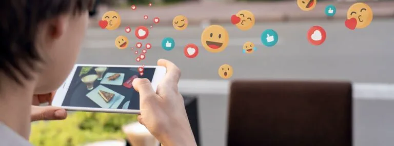 Person holding smartphone showing food photos with social media emoji reactions floating above.