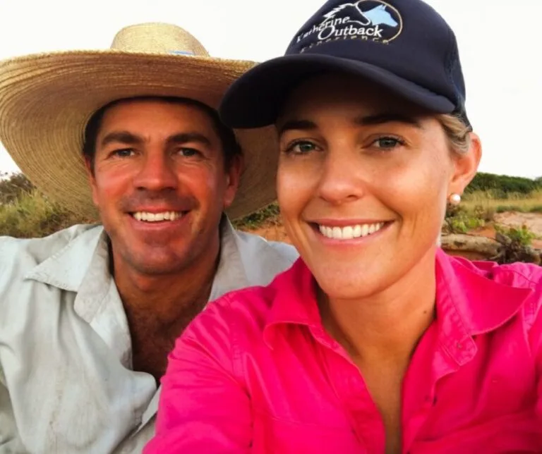 Two people smiling at camera in outback setting, man in straw hat and woman in pink shirt.