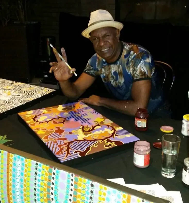 Aboriginal artist displaying colourful artwork at workshop table with paint supplies.