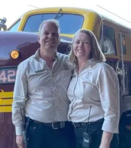 Fab and Liz in branded shirts standing in front of vintage train carriages.