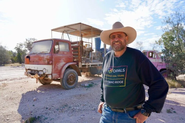 Tourism operator in cowboy hat stands in outback setting with vintage red truck behind.