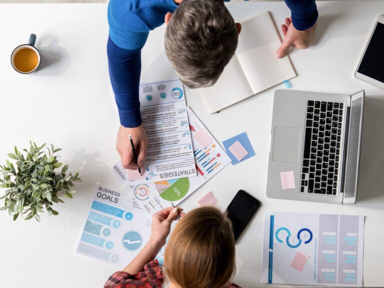 Two people review business strategy documents and charts at white desk workspace.