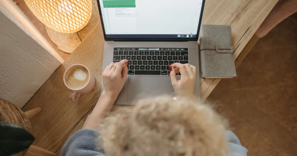 Overhead view of hands typing website content on a laptop, with a latte and notebook on a warm wooden desk.